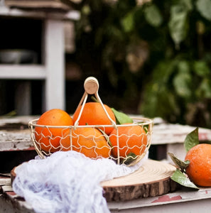 Outdoor table setting with large leafy shrubs in the background. On the table is a cream metal basket filled with bright, ripe oranges.