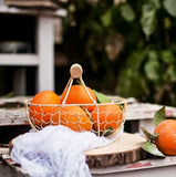 Outdoor table setting with large leafy shrubs in the background. On the table is a cream metal basket filled with bright, ripe oranges.