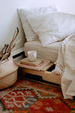 Boho bedroom with a neutral comfortable looking bed, a stack of books and a coffee mug, a colourful patterned rug, and a natural seagrass basket beside the bed.