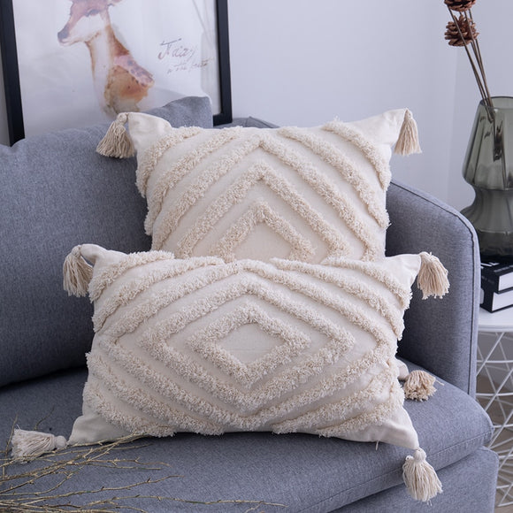 Two cream pillows with a textured diamond in the centre and incrementally larger diamonds radiating toward the outer edges are sitting on a grey couch. The pillows have a cream tassel on each corner. 