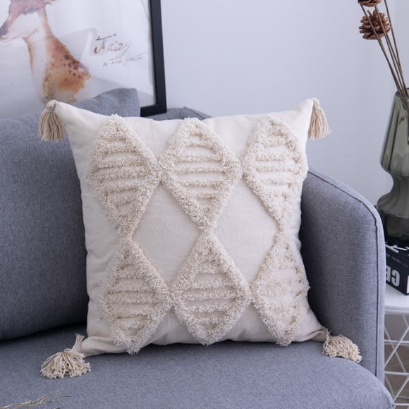 18 by 18 cream pillow with textured diamonds creating an argyle pattern, with alternating cream fabric diamonds and textured diamonds with textured horizontal lines inside; pillow corners have cream tassels. Pillow is placed in the corner of a grey couch.