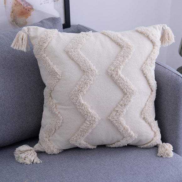 18 by 18 inch cream pillow with textured zig zags and cream tassels in each corner is placed in the corner of a grey couch.