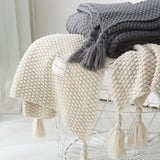 Cream chunky blanket is draped on a white coffee table with a dark grey chunky knit throw blanket folded on top. Shows chunky knit and large tassels up close.