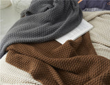 Cream, brown, and grey chunky knit throw blankets are layered on a comfortable bed with an open book.