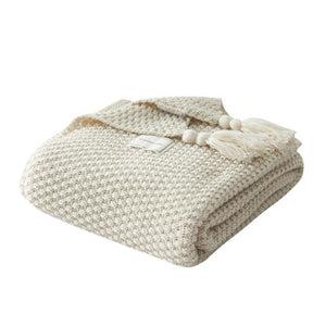 Cream chunky blanket is draped on a white coffee table with a dark grey chunky knit throw blanket folded on top. Shows chunky knit and large tassels up close.