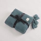 Folded blue/grey chunky knit blanket with large tassels. Blanket is tied with a large ribbon.