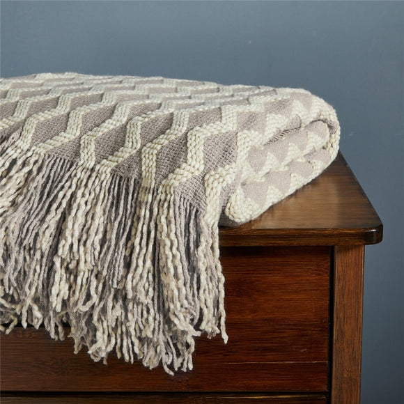 Beige blanket with cream zig zag knit pattern is folded atop of a wooden dresser with the full fringe edge hanging over the edge of the dresser.
