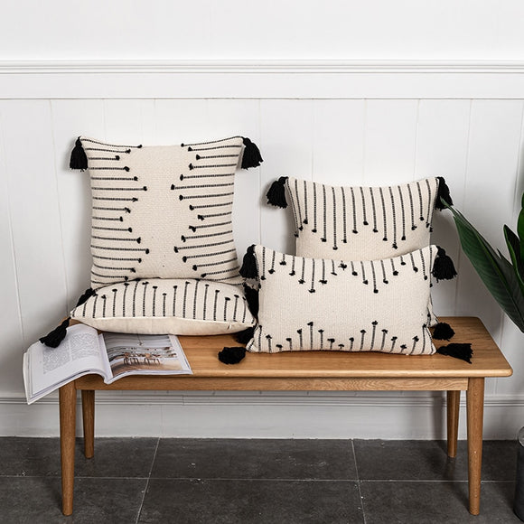 Wooden bench with 4 cream pillows on it. Pillows are woven with black tufts to create a geometric design. Pillow corners have black tassels.